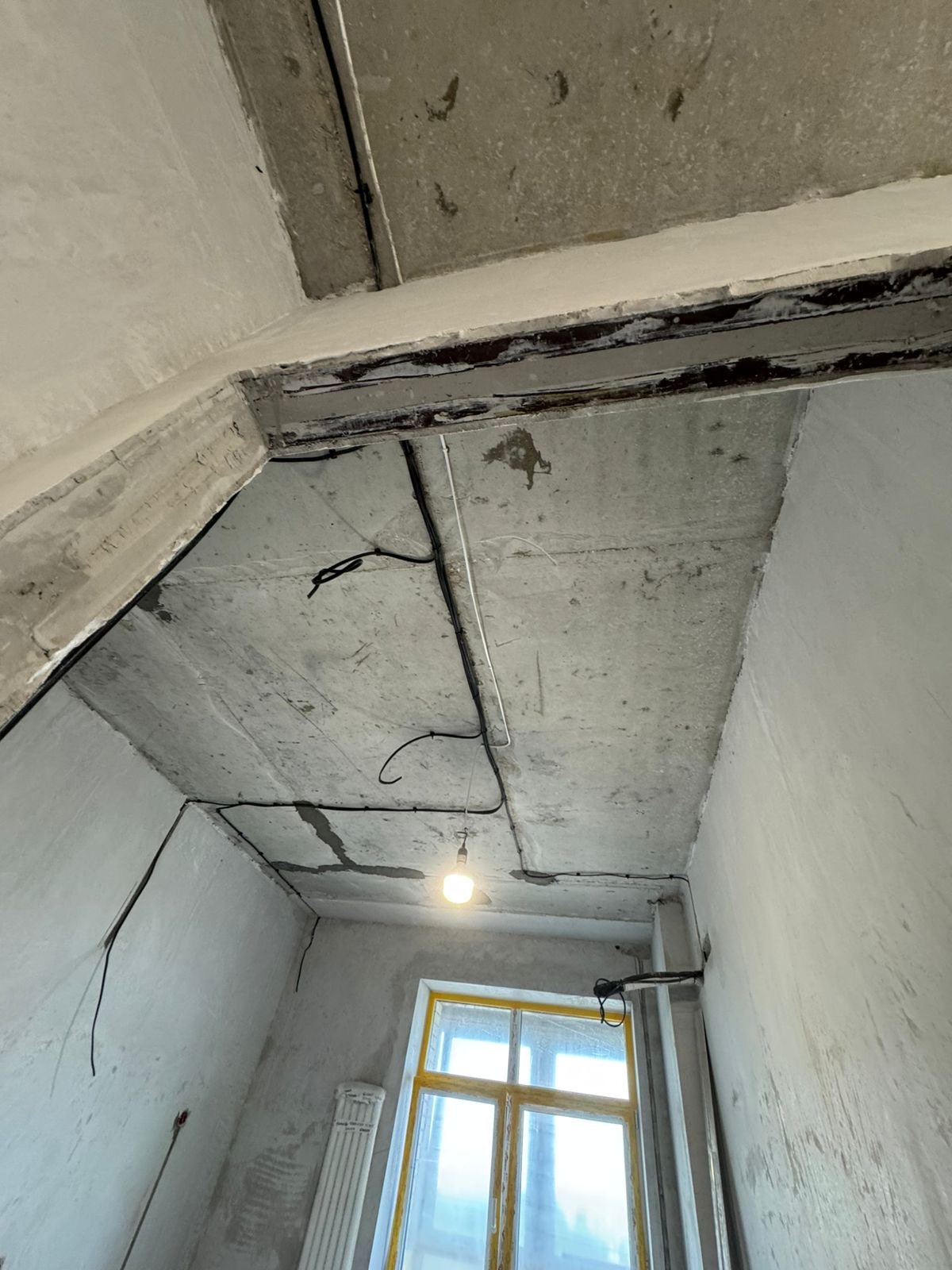 Электропроводка под потолком. Вид снизу. Бетонный потолок, провода, лампочка, окно.
