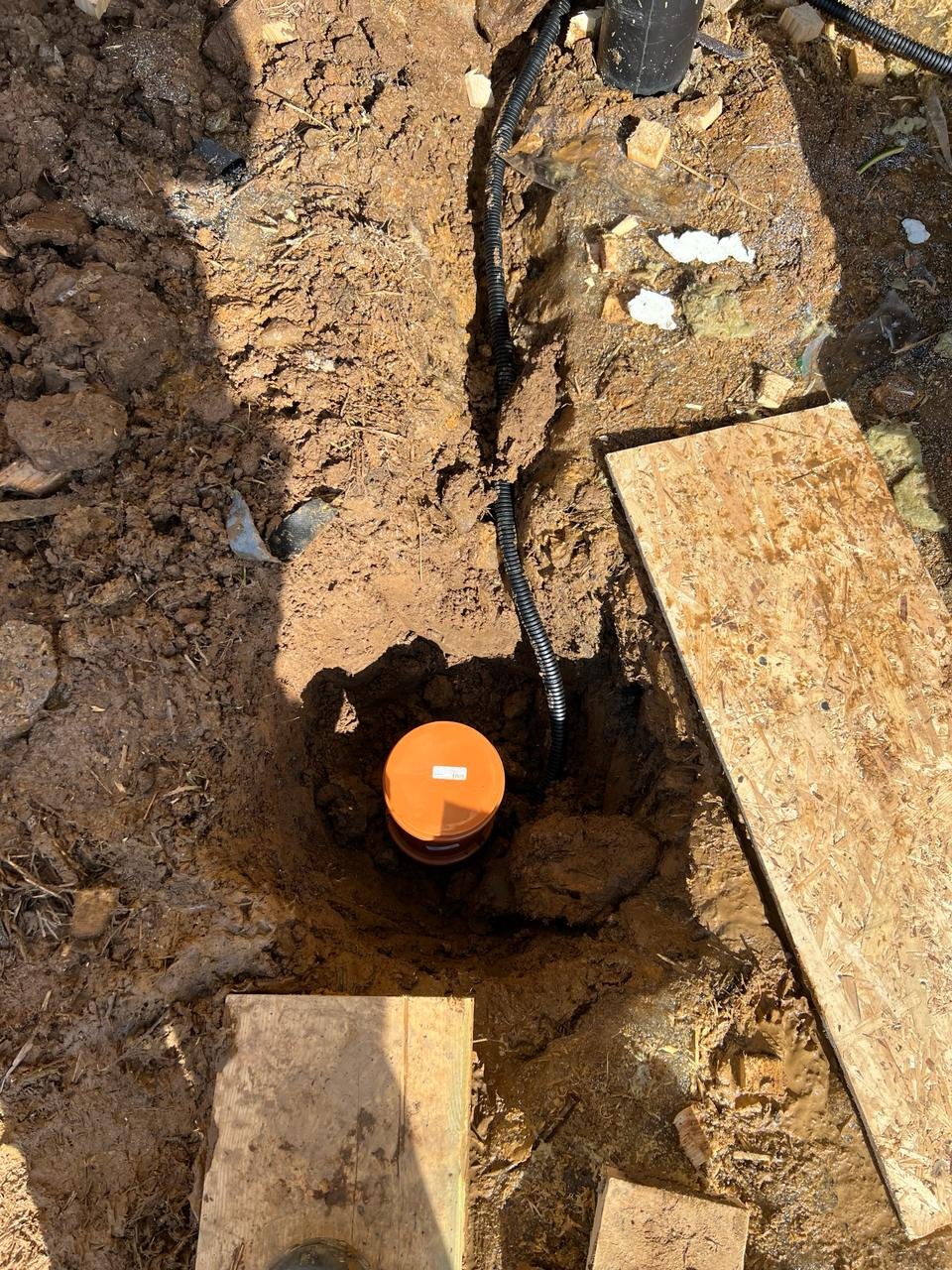 Заземление в траншее: оранжевая труба, провода в гофре, земля, доски.
